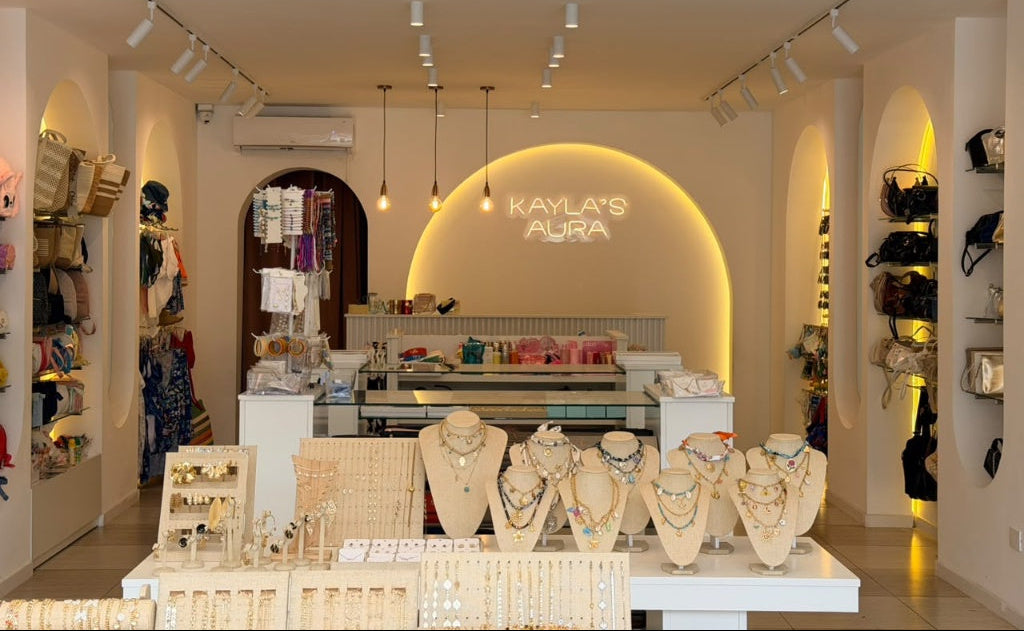 A jewelry boutique interior with multiple display busts showcasing necklaces and rings on tables, shelving with bags and accessories along the walls, and a back wall illuminated with a glowing 'KAYLA'S AURA' sign.